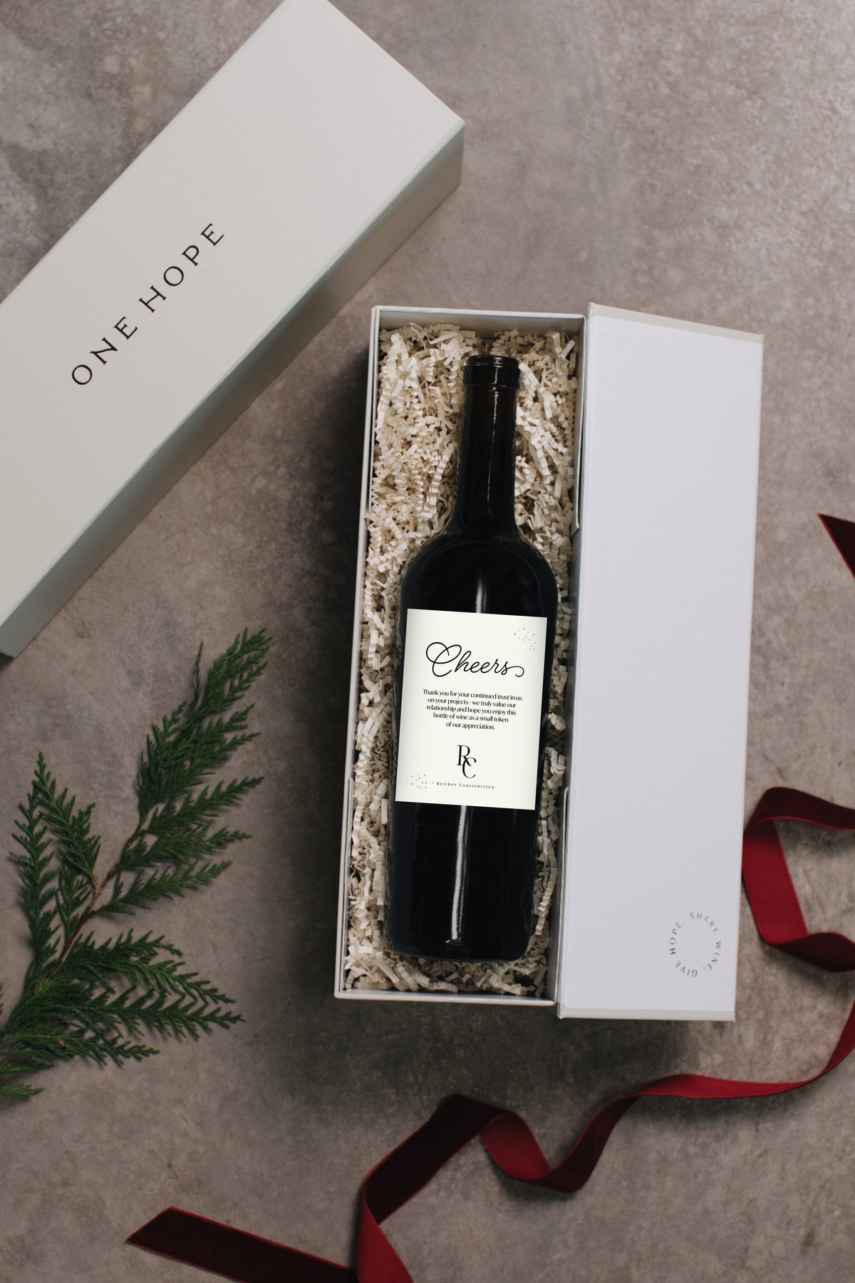 A wine bottle presented in a gift box with shredded filler, alongside the matching closed box lid and a red ribbon, arranged on a textured gray surface with a green sprig.